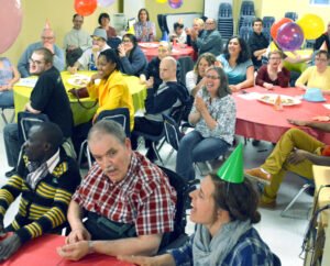 A diverse group of individuals at a celebration event, seated at tables with colorful tablecloths. Many are wearing party hats and smiling, while balloons are visible in the background. This gathering supports L’arche Disability Transformations.