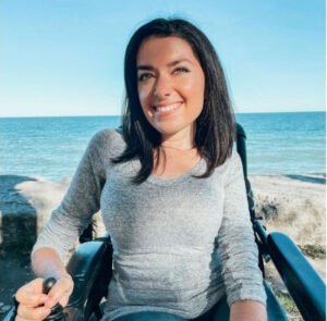 A young woman with dark hair smiling while sitting in a wheelchair by a lakeside. The background shows a clear blue sky and water, with rocky terrain in the foreground, illustrating accessibility issues while flying.
