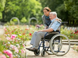 A caregiver assisting an elderly man in a wheelchair, surrounded by blooming roses in a garden. The caregiver is smiling and leaning slightly over the man, who appears content and engaged with the scenery, reflecting themes of Assistive Health Care.