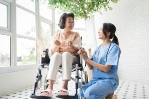A healthcare worker in blue scrubs assists an elderly woman in a wheelchair, both smiling in a well-lit room with large windows and greenery. This scene represents Winnipeg home care healthcare.