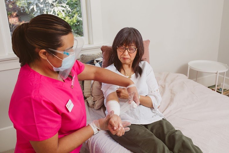 A healthcare worker in pink scrubs applying a bandage to the arm of a patient seated on a bed in a bright room. The patient appears calm and is wearing glasses.