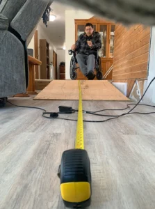 A person in a wheelchair on a wooden ramp, measuring the length of the ramp with a tape measure placed on the floor. The setting appears to be inside a home.