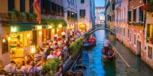 A picturesque canal in Venice lined with lively restaurants and diners enjoying outdoor seating. A gondola navigates through the water, surrounded by historic buildings with green shutters. Expedia: Accessible Travel.