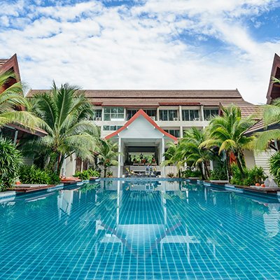 An outdoor swimming pool surrounded by tropical palm trees and greenery, with a modern building in the background under a partly cloudy sky. The scene conveys a tranquil resort atmosphere, supporting the theme of 'Access Now: Pin-Pointing Accessible PlaCES around the world'.