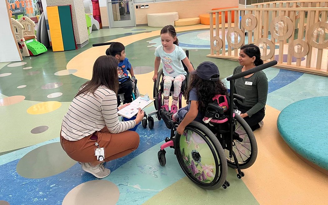A group of children in wheelchairs and two adults interact in a colorful play area. One child smiles while talking to an adult, and another child listens. The setting features playful designs on the floor, emphasizing the Rehab Centre for Children Program.