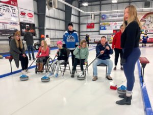 A group of people participating in a wheelchair curling event in Steinbach. Some sit on chairs while curling stones are positioned on the ice. Participants include youth and adults, showcasing the inclusive nature of the sport.