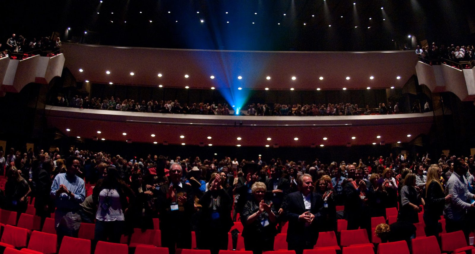 An audience stands and applauds in a theater, with a spotlight illuminating the stage. The seating area features red chairs, and attendees are clapping in appreciation. This image showcases the atmosphere of a performance at Centennial Concert Hall.