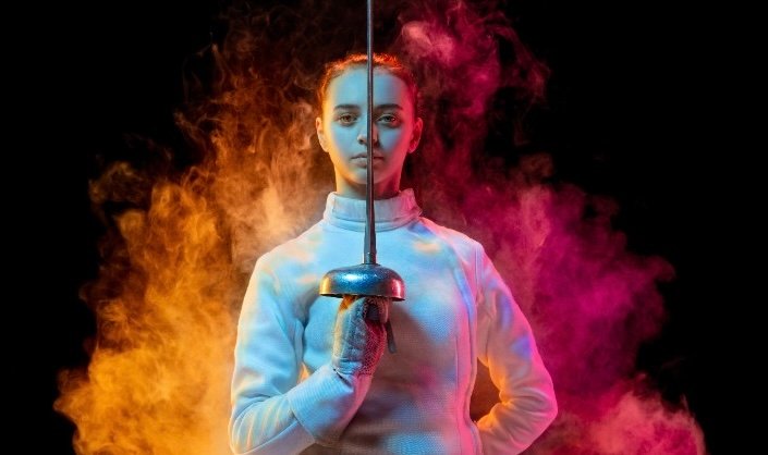 A fencer in a white uniform stands confidently, holding a foil sword upright. Colorful smoke in shades of orange and purple surrounds her against a black background.