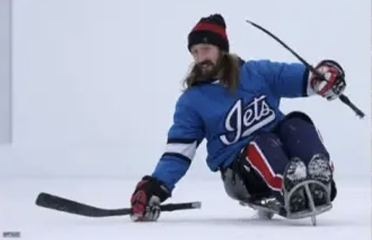 A man with long hair wearing a blue hockey jersey and snow pants is playing sled hockey on ice. He is holding a hockey stick and is in a dynamic pose, sliding past a black hockey puck.