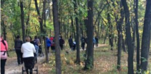 A group of people walking along a winding path in a forested area with trees displaying autumn foliage. Some individuals are pushing strollers. The scene illustrates outdoor activity in a natural setting.