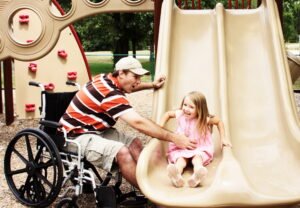 A man in a wheelchair assists a young girl as she prepares to slide down a playground slide. They are smiling, surrounded by play equipment in a park setting, highlighting engaging moments between them.
