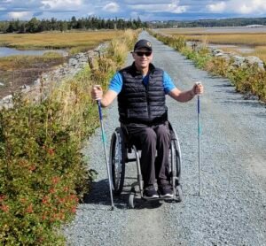 A person in a wheelchair on a gravel path surrounded by grass and water, using walking poles. The setting is bright, with trees and a cloudy sky in the background, showcasing the Trans Canada Trail All-Persons Trail.