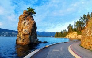 A scenic waterfront path winding along a rocky shoreline, with a large rock formation topped by a small tree emerging from the water. In the background, mountains and a city skyline are visible under a blue sky.