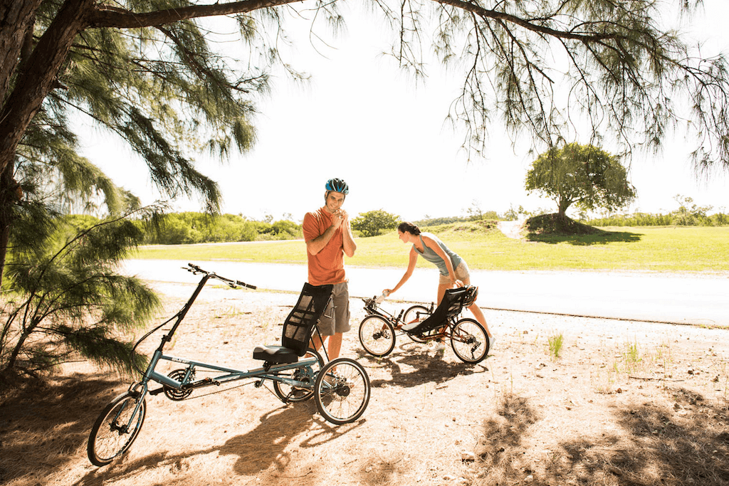 Two individuals near recumbent bicycles in an outdoor setting, surrounded by trees and grass. One person appears to be inspecting a bike while the other stands nearby in casual attire. This image relates to 'Recumbent Cycling: Complete Beginners Guide'.