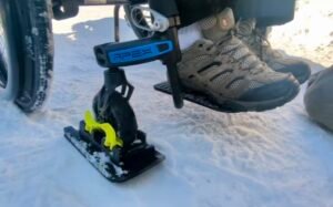 Close-up view of a wheelchair equipped with a yellow snow sled attachment on a snowy surface. The user's shoes are visible, showing traction tread suitable for outdoor conditions. This image relates to Accessible Sport - Events List.