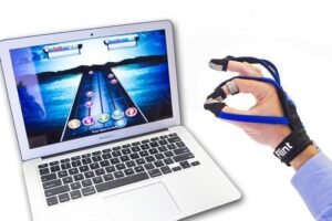 A laptop displaying a colorful rhythm game interface with notes on a highway, alongside a hand wearing a musical hand therapy device that enhances finger mobility and coordination.