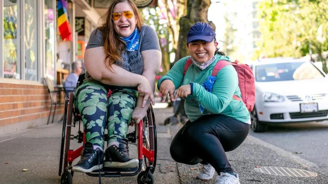 Two women making a victory sign. One is sitting on a wheelchair while the other is squatting on the ground. Both are smiling brightly.