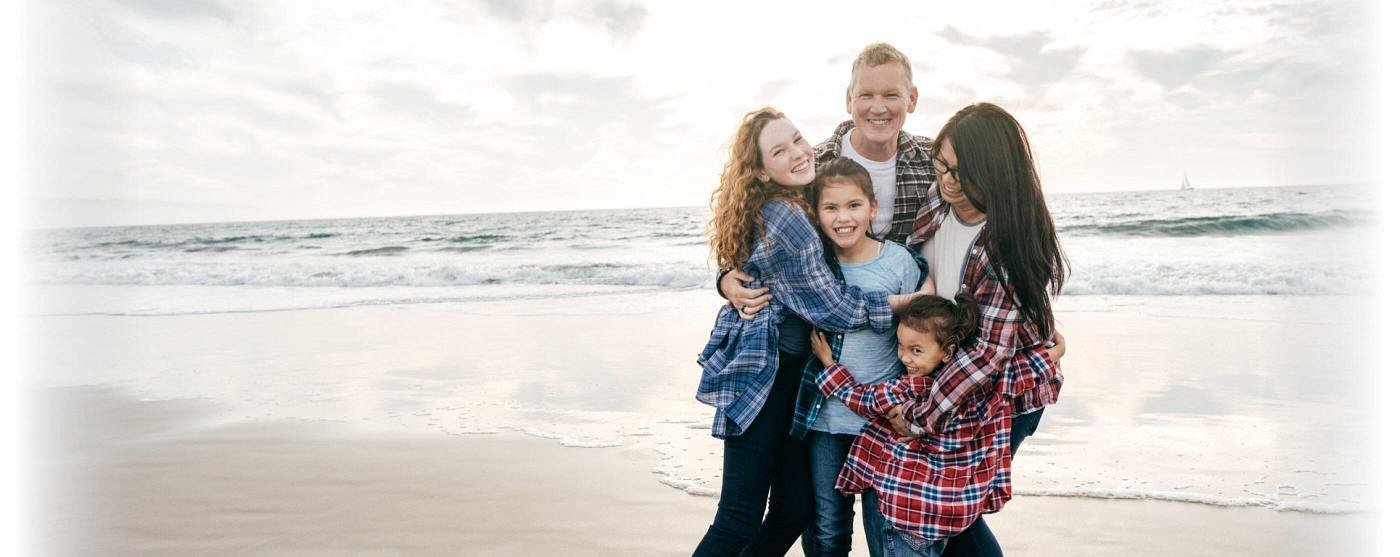 A family of five standing on the shore of the sea while hugging each other and smiling brightly.