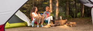 Two women caping in the forest, sitting Infront of the campfire while sipping coffee and smiling cheerfully.