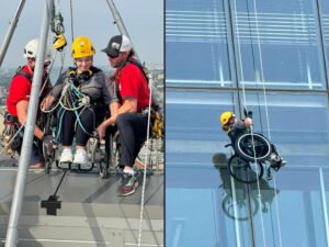 Two men helping a woman in a wheelchair as she gears up to rappel down a building.