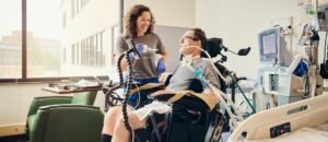 Man seated with medical equipment attached, while a smiling woman conducts a checkup.