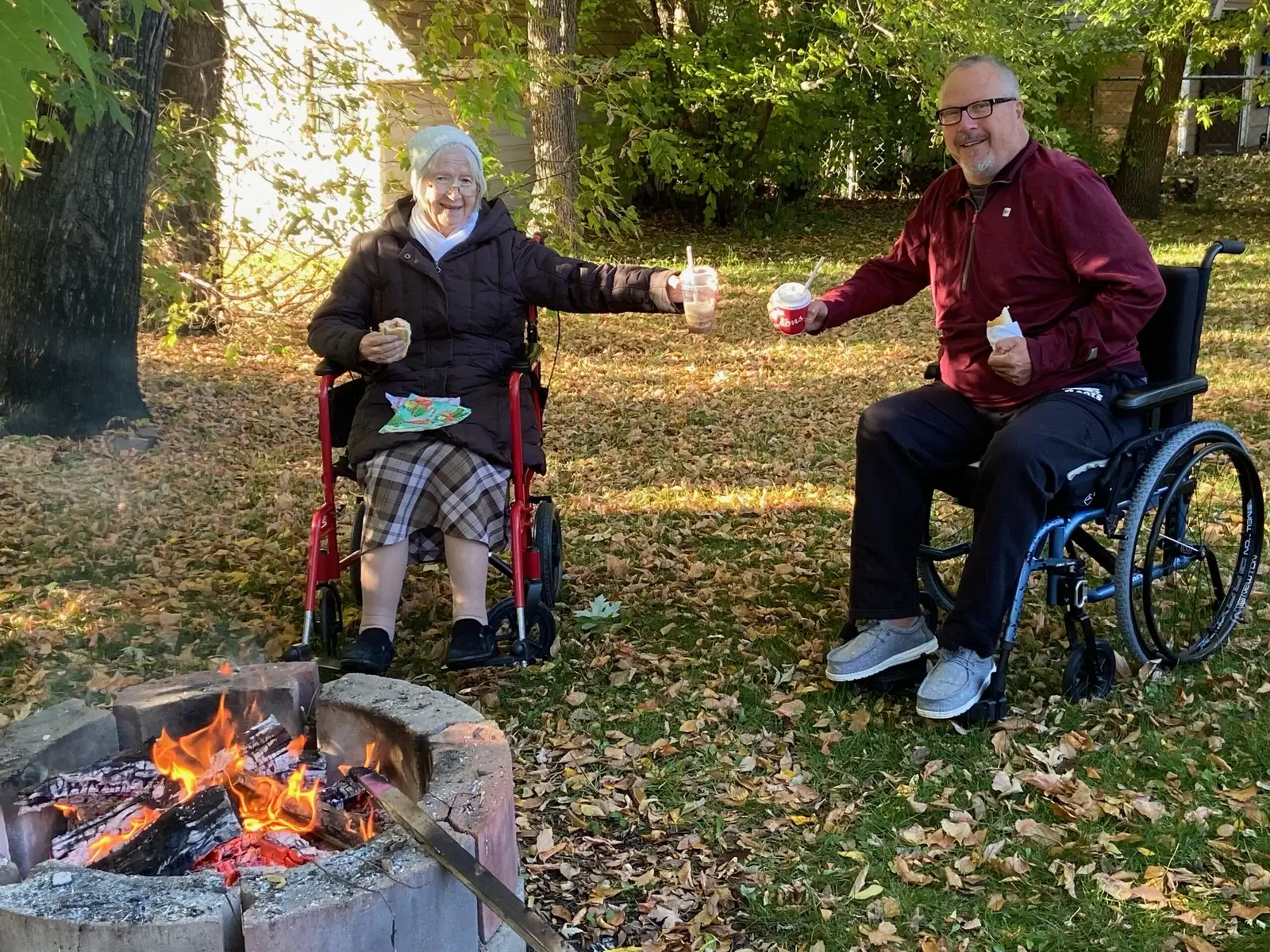 Elderly woman and middle-aged man in wheelchairs holding food by a charcoal grill in a lush garden.