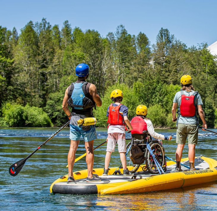 A family of four navigating a river on a raft, one of them is in wheelchair, smiling and enjoying their outdoor adventure.
