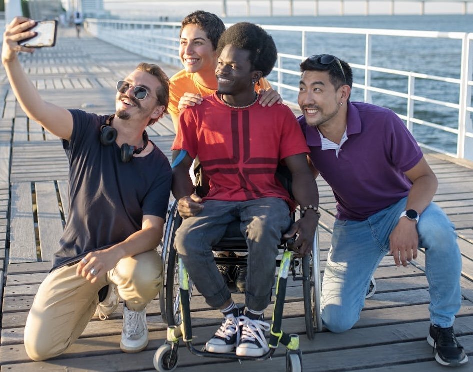 Group of four diverse friends, including a man in a wheelchair, smiling and taking a selfie on a boardwalk by the water on a sunny day.