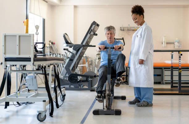 A woman in a white lab coat is standing by an old lady who is using a stationary bike in a clinical.