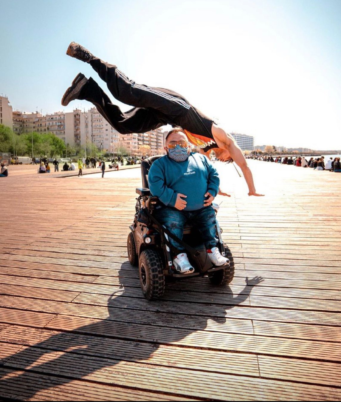 A man in a wheelchair and another man behind him executes a handstand on a wooden board, demonstrating remarkable skill and athleticism.