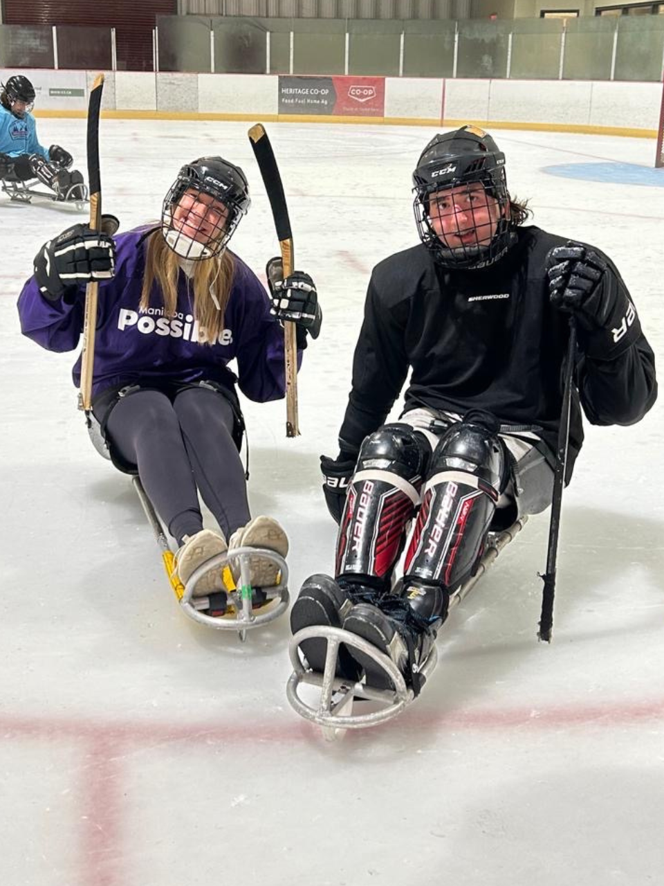 Two people on skates sitting on an ice rink, surrounded by a smooth, reflective surface of ice.