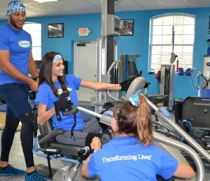 A man and a woman wearing blue shirts are actively pedaling on stationary bikes during a workout session while another girl sitting by side