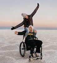 Two women in wheelchair on a bright salt flat, surrounded by a serene, open sky.