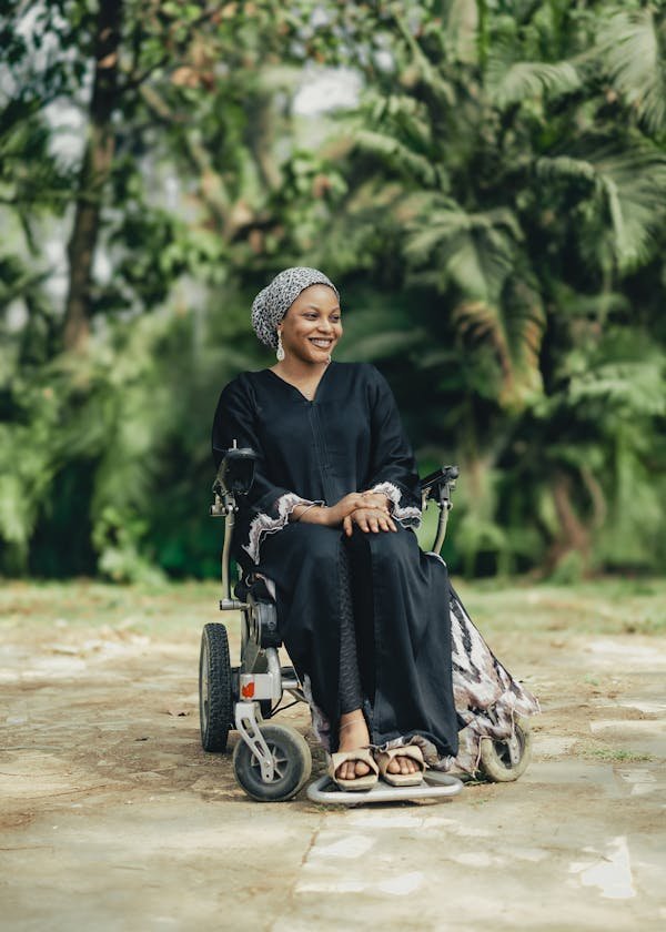 A woman dressed in black sitting in a wheelchair is outside on the walk approach to her home, sitting with a smile on her face