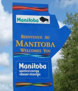 A large sign welcoming visitors to Manitoba, featuring the text 'Bienvenue Au Manitoba' and 'Manitoba Welcomes You.' It includes images of the region's silhouette and a bison logo.