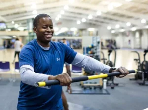 A man smiling while using a resistance training machine in a gym. He is wearing a blue athletic shirt and is engaged in a workout, with exercise equipment in the background.