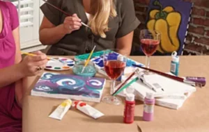 Two people engaged in painting at a table, with paintbrushes, palettes, and canvases. There are glasses of red wine and various paint tubes scattered on the surface. A colorful artwork is visible in the background.