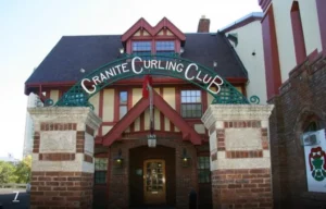 Exterior view of the Granite Curling Club, featuring a red and beige facade with a green sign above the entrance. The image captures the club's architectural details, characteristic of Winnipeg. A notable tourist attraction.