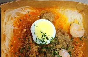 A bowl of noodles topped with ground meat, a poached egg, shrimp, and garnished with chopped herbs. The dish has a savory orange sauce and is presented in a brown takeout container.