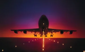 A silhouette of an airplane landing against a vibrant sunset with shades of purple and orange. Lights are visible on the runway, creating a guiding path for the aircraft.