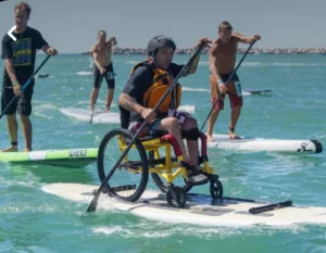 Individuals participating in a paddleboarding event on the ocean. A person in a wheelchair is using a modified paddleboard, while other participants paddle nearby.