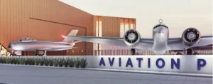 Two vintage aircraft displayed at the entrance of an aviation park, with a modern building in the background. The foreground features a sign with the word "AVIATION" prominently displayed.