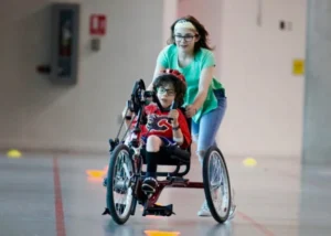 A young woman guiding a child in a specially designed handcycle, both navigating an indoor track with colored cones. The child wears glasses and a helmet, while the woman is dressed in a green shirt.