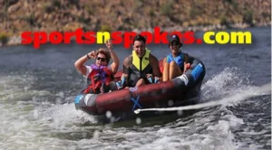 Three people enjoying a ride on a rubber inflatable boat on water, surrounded by hills. Two are wearing life jackets, and all are smiling. The water is choppy from the boat's movement.