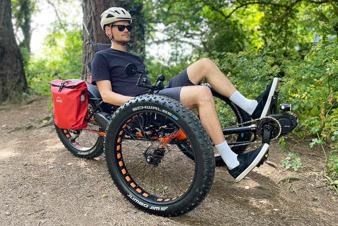 A cyclist using an adaptive three wheel cycle is pending through a forest of greenery on a dirt trail