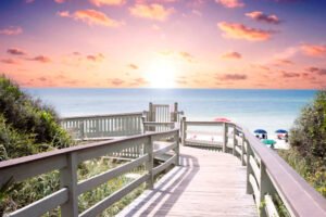 Image shows the sun setting over the ocean and there’s a beautifully designed accessible ramp with railings going down to the shore