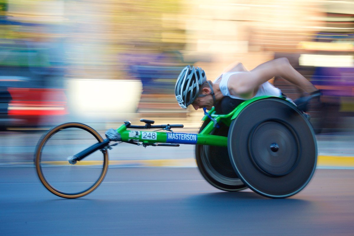Disabled athlete is hunched over and pushing the rear wheels of his three wheeler racing cycle, and the image is blurred in the background indicating he’s going fast