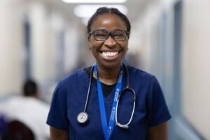 Nurse with stethoscope around her neck, and a big smile is in the standing in the corner of the hospital