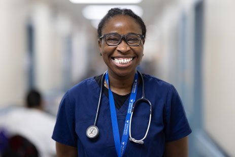 Nurse with stethoscope around her neck, and a big smile is in the standing in the corner of the hospital