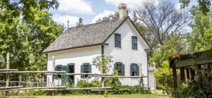 Shows an old white flatboard house sat in the trees with some rustic fencing around it